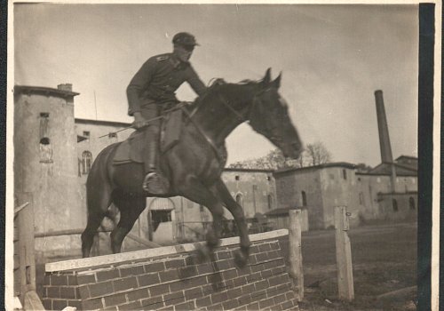 Wymiary: wysokość - 9 cm, szerokość - 12 cm; Materiał: papier fotograficzny; Technika: fotograficzna; Pochodzenie: Żory, G&oacute;rny Śląsk; Datowanie: lata 1922-1939; Autor: brak danych 
Fotografia czarno-biała przedstawiająca żołnierza (II Dywizjonu 23 Pułku Artylerii Lekkiej w Żorach) na koniu pokonującego przeszkodę na placu ćwiczeń. Na drugim planie widoczne zabudowania garnizonowe.